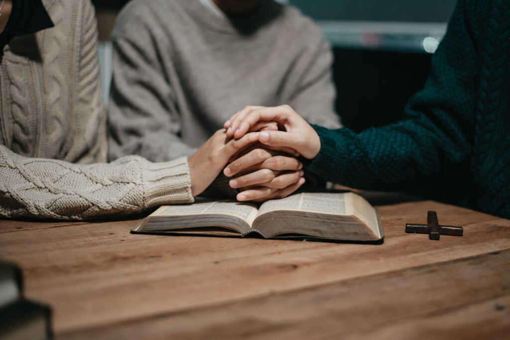 Family holding hands in prayer around an open Bible on a wooden table
